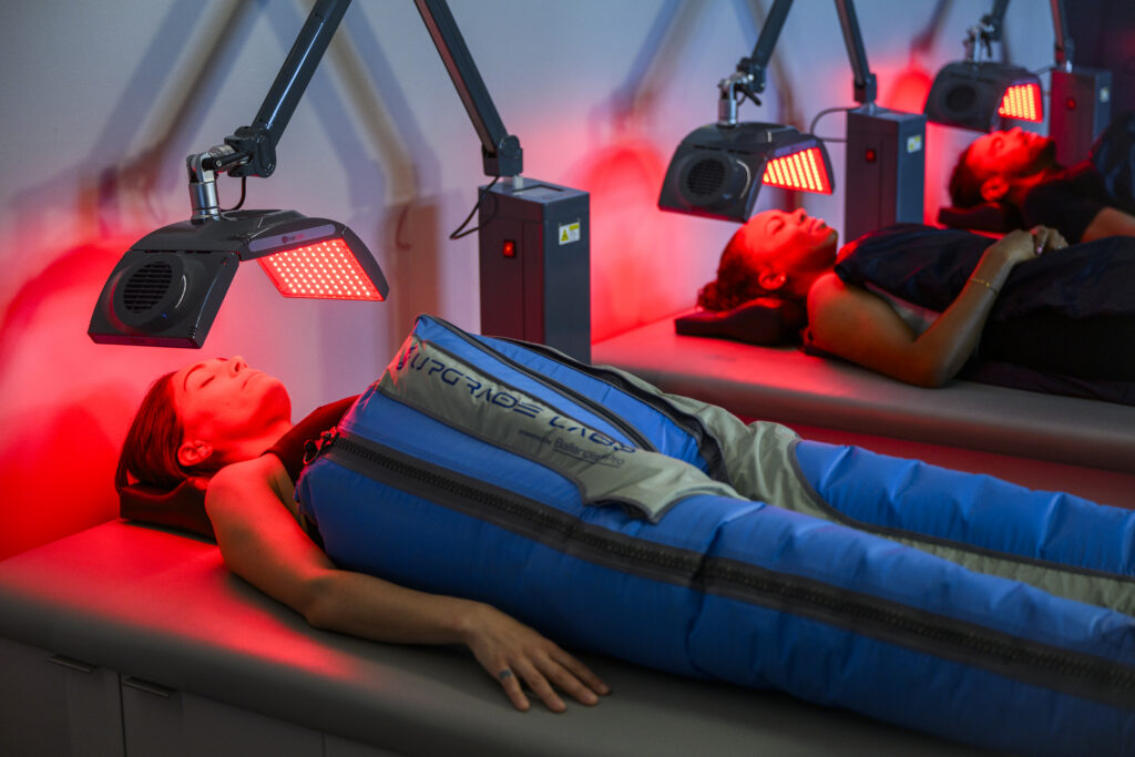 Woman lying down in Big Squeeze compression pants while two people receive PEMF therapy in the background.