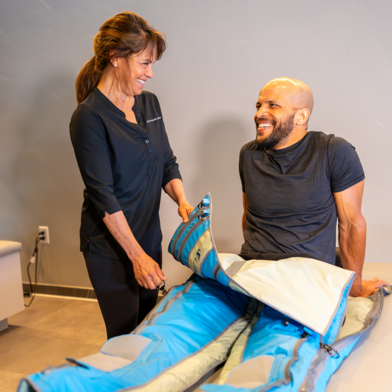 A woman in a black Upgrade Labs uniform is assisting a man in black athletic wear as he get out of the Big Squeeze™ at Upgrade Labs, both are looking at each other and smiling.