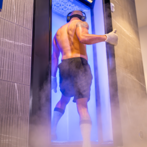 A man getting into cryotherapy machine at A shirtless man wearing athletic shorts, ear-protecting headphones, gloves, and high socks is entering a cryotherapy machine at Upgrade Labs.